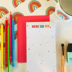 Op de afbeelding: Een wit notitieboekje met de tekst "MERK ICH MIR" in kleurrijke letters en een klein hartje. Het notitieboekje ligt op een roze envelop en een witte achtergrond met regenboogpatronen. Er zijn kleurpotloden en een markeerstift aan de linkerkant van de afbeelding.
