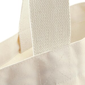May include: A close-up view of an off-white canvas tote bag featuring thick, woven fabric handles securely stitched to the main body with a reinforced box-and-cross pattern. The material has a visible, textured weave characteristic of natural cotton.