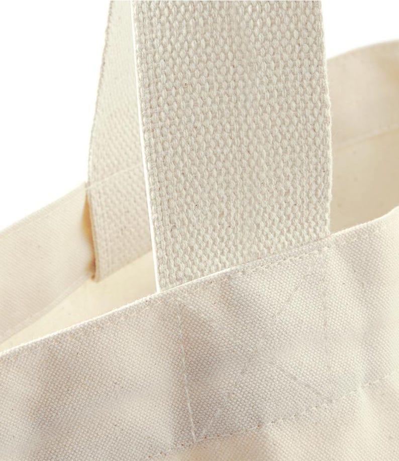 May include: A close-up view of an off-white canvas tote bag featuring thick, woven fabric handles securely stitched to the main body with a reinforced box-and-cross pattern. The material has a visible, textured weave characteristic of natural cotton.