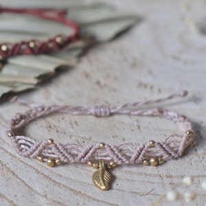 May include: Two braided bracelets with gold beads and leaf charms. The bracelet on the left is a dark red color and the bracelet on the right is a light pink color.
