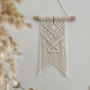Puede incluir: Colgante de pared de macramé blanco con un patrón geométrico, colgado de una barra de madera. Una guirnalda de luna y estrellas doradas cuelga debajo del macramé.