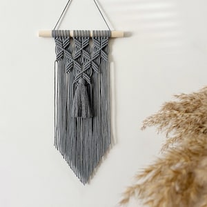 May include: Gray macrame wall hanging with a wooden dowel and a tassel. The wall hanging is in a diamond pattern.