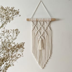 May include: A white macrame wall hanging with a geometric pattern and a tassel. The hanging is attached to a wooden dowel rod.