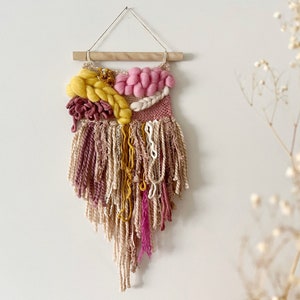 May include: A pink, yellow and cream woven wall hanging with a wooden dowel at the top. The hanging has a fringe of yarn and is decorated with pom-poms.