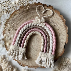 May include: A pink and cream rainbow macrame wall hanging with a heart-shaped loop at the top and a small bow. The rainbow is made with a variety of textures and colours, including pink, cream, and gold.