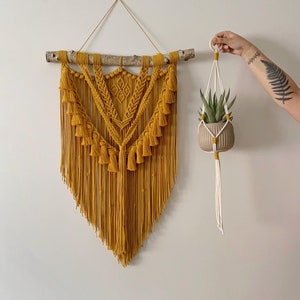 May include: A mustard yellow macrame wall hanging with tassels, featuring a geometric pattern, is hung on a branch. A white macrame plant hanger with a small potted plant is hanging on the right side of the image.