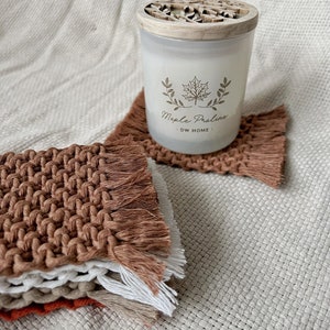 May include: A white candle with a wooden lid featuring a leaf design. The candle is labeled "Maple Praline - DW HOME". The candle is sitting on a brown crocheted coaster with fringe.