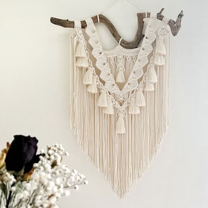 May include: A white macrame wall hanging with a natural wood branch at the top. The macrame features a geometric pattern with tassels and fringe.