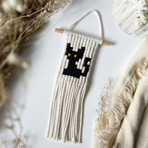 May include: A black cat with yellow eyes is woven into a white macrame wall hanging with fringe.
