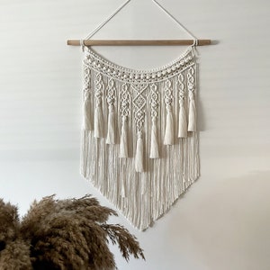 Puede incluir: Un tapiz de macramé blanco con una barra de madera en la parte superior. El tapiz presenta un patrón geométrico con borlas en la parte inferior.
