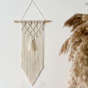 Könnte beinhalten: Ein weißer Makramee-Wandbehang mit einem Holzstab oben und einem Quasten in der Mitte. Der Behang ist im Diamantenmuster gehalten und hat unten einen Fransen.