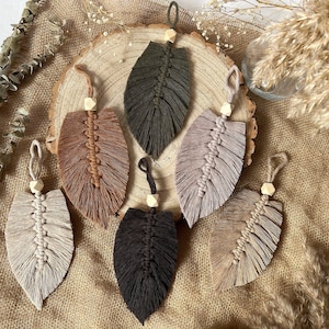 May include: Six macrame leaf wall hangings with wooden beads. The leaves are in various colors, including brown, beige, black, and green.