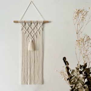 May include: A white macrame wall hanging with a geometric pattern and a tassel. The hanging is attached to a wooden dowel rod.