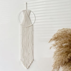 May include: White macrame wall hanging with a circular design and a long, beaded fringe. The hanging is against a white wall.