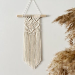 Puede incluir: Un tapiz de macramé blanco con un patrón geométrico y flecos. El tapiz está sujeto a una varilla de madera.