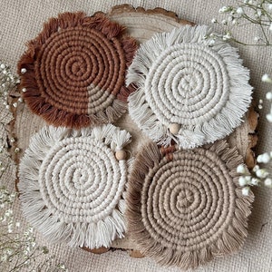 May include: Four handmade macrame coasters with fringe edges. The coasters are in shades of brown, beige, and white. Each coaster has a wooden bead in the center.