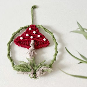 May include: A handmade macramé mushroom wall hanging. The mushroom cap is red with white dots, and the stem is beige. The mushroom is surrounded by a green macramé circle and green leaves.
