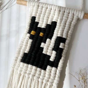 May include: A white macrame wall hanging with a black and yellow cat design. The hanging has a wooden dowel at the top and long white fringe at the bottom.