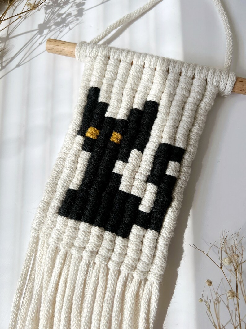 May include: A white macrame wall hanging with a black and yellow cat design. The hanging has a wooden dowel at the top and long white fringe at the bottom.