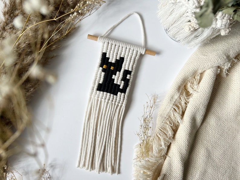 May include: A black cat with yellow eyes is woven into a white macrame wall hanging with fringe.
