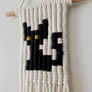 May include: A black and white macrame wall hanging with a black cat design and yellow eyes. The hanging has a wooden dowel at the top and long white fringe at the bottom.