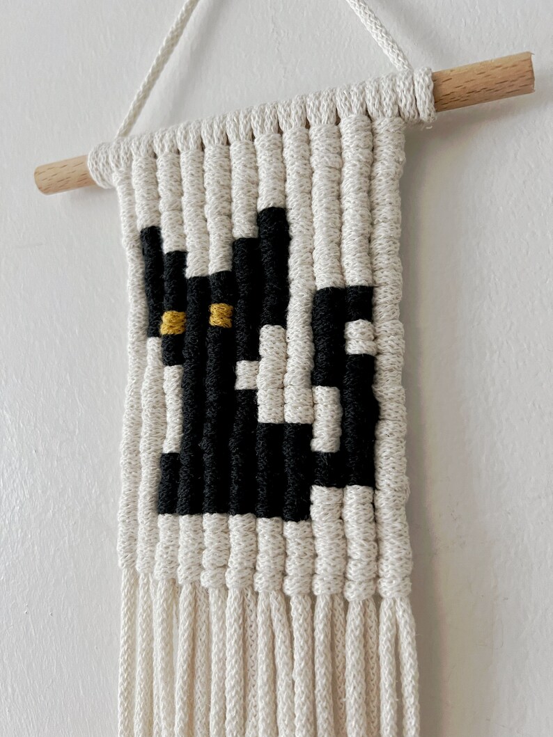 May include: A black and white macrame wall hanging with a black cat design and yellow eyes. The hanging has a wooden dowel at the top and long white fringe at the bottom.