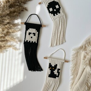 May include: Three handmade macrame wall hangings with Halloween themes. The first is a white ghost on a black background, the second is a black skull on a white background, and the third is a black cat on a white background.