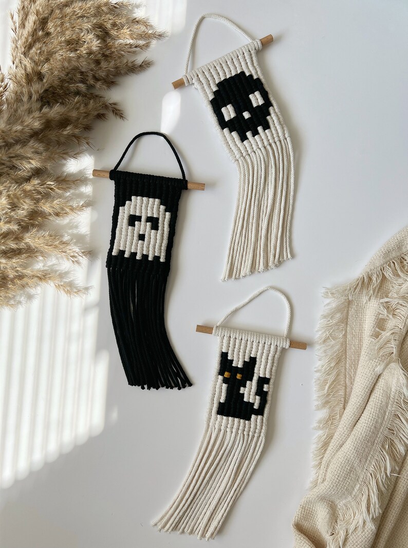 May include: Three handmade macrame wall hangings with Halloween themes. The first is a white ghost on a black background, the second is a black skull on a white background, and the third is a black cat on a white background.
