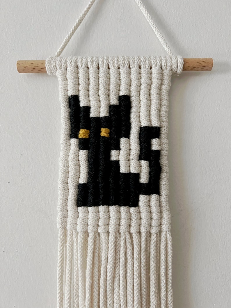 May include: A black and white macrame wall hanging with a cat design. The cat has yellow eyes and is surrounded by a white fringe.