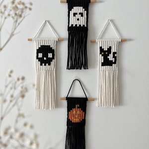 May include: Four Halloween-themed wall hangings with black and white yarn. The hangings feature a ghost, a skull, a black cat, and a pumpkin. Each hanging has a wooden dowel at the top and a fringe of yarn at the bottom.
