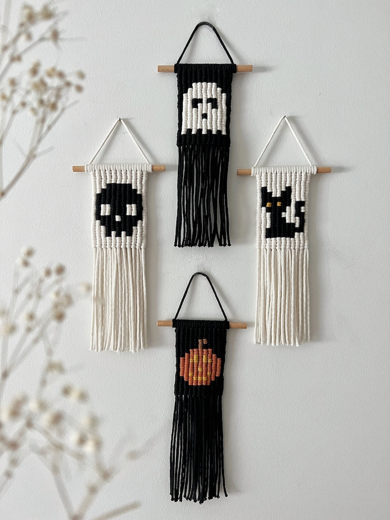 May include: Four Halloween-themed wall hangings with black and white yarn. The hangings feature a ghost, a skull, a black cat, and a pumpkin. Each hanging has a wooden dowel at the top and a fringe of yarn at the bottom.