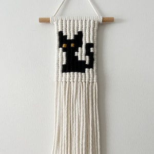 May include: A black cat macrame wall hanging with white yarn and a wooden dowel at the top. The cat has yellow eyes.