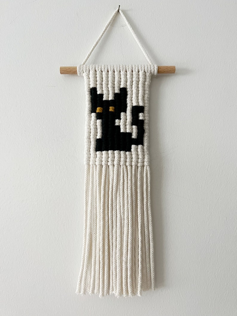 May include: A black cat macrame wall hanging with white yarn and a wooden dowel at the top. The cat has yellow eyes.