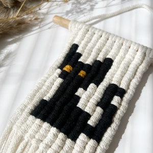 May include: A black and white macrame wall hanging with a black cat design and yellow eyes. The hanging is made with natural cotton cord and has a wooden dowel at the top.