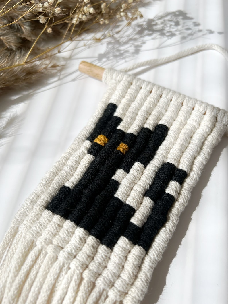 May include: A black and white macrame wall hanging with a black cat design and yellow eyes. The hanging is made with natural cotton cord and has a wooden dowel at the top.