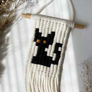 May include: A black and white macrame wall hanging with a black cat design and yellow eyes. The hanging has a wooden dowel at the top and a fringe at the bottom.