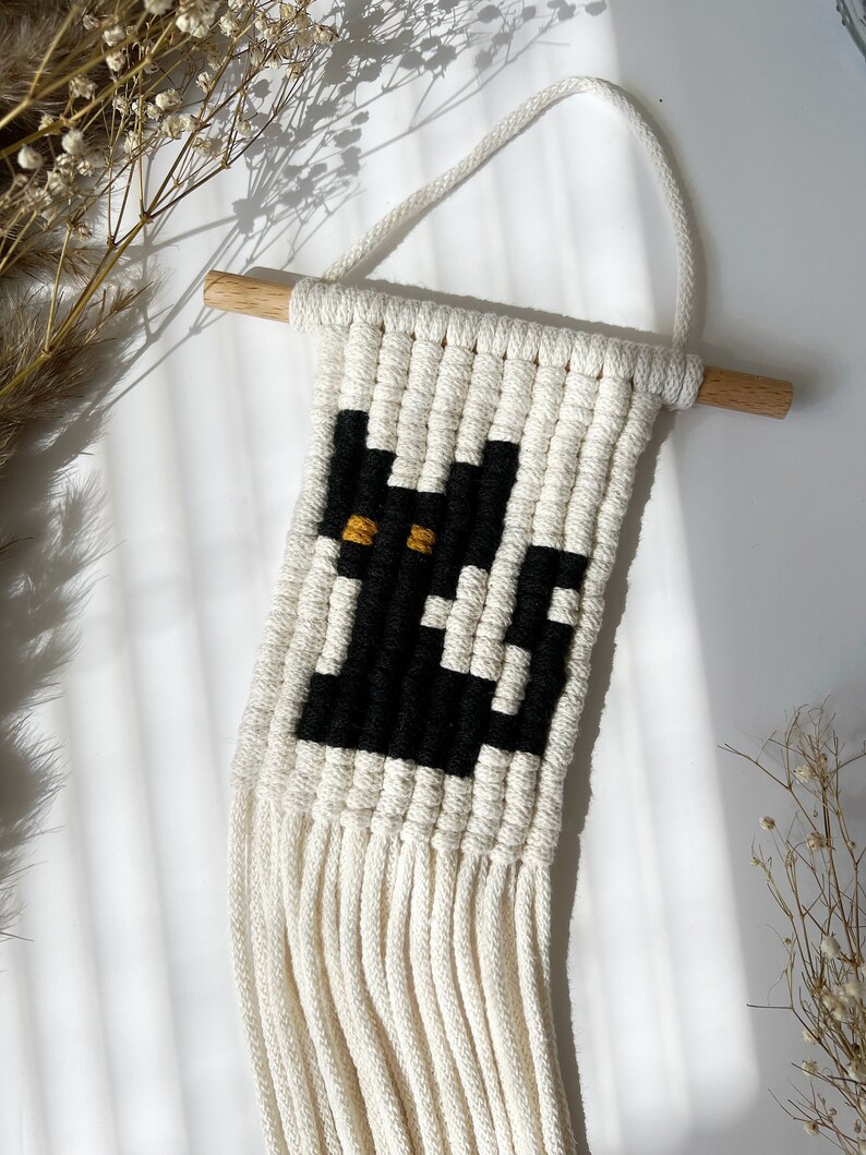 May include: A black and white macrame wall hanging with a black cat design and yellow eyes. The hanging has a wooden dowel at the top and a fringe at the bottom.