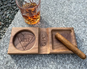 Puede incluir: Un cenicero de madera para cigarros con un posavasos redondo para un vaso y un reposo rectangular para un cigarro. El cenicero presenta el emblema de la Policía Estatal de Massachusetts y las iniciales "GPC". Un vaso de líquido ámbar está cerca.