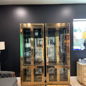 May include: Two tall, gold-toned glass-front cabinets with mirrored backs and geometric patterned shelves. The cabinets are standing on a patterned rug in a room with a black wall.