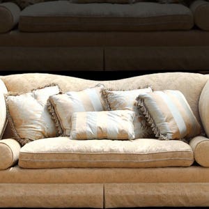 May include: A large, beige, tufted sofa with rolled arms and a decorative skirt. The sofa has five throw pillows, two of which are solid beige, two are striped beige and white, and one is solid white.
