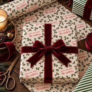 May include: A rectangular gift wrapped in white paper featuring a repeating pattern of green holly leaves and red berries, with the text "with love from the walkers" in red script. A deep red velvet ribbon is tied in a bow around the center.