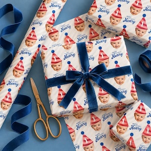May include: Wrapping paper featuring a repeating pattern of a baby wearing a red and white striped party hat with the number one, accompanied by the text Happy Birthday Henry, accented by a dark blue velvet ribbon and gold scissors.