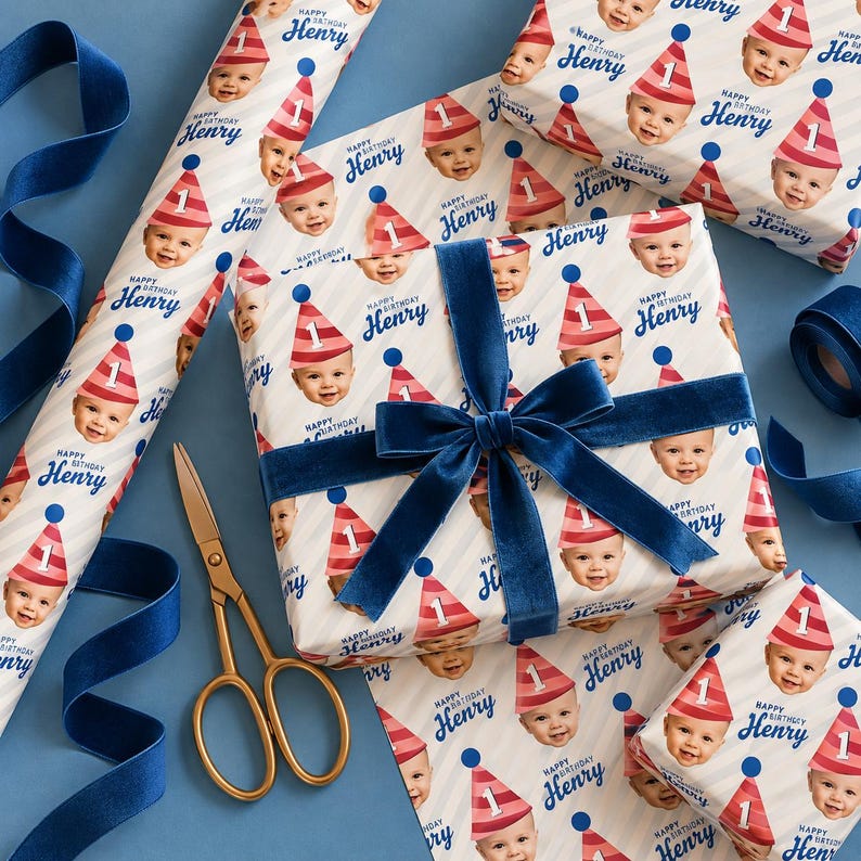 May include: Wrapping paper featuring a repeating pattern of a baby wearing a red and white striped party hat with the number one, accompanied by the text Happy Birthday Henry, accented by a dark blue velvet ribbon and gold scissors.