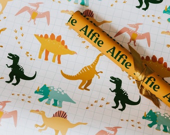 May include: Wrapping paper featuring a dinosaur pattern in shades of yellow, green, and pink, with a grid background. A gold party cracker with the name "Alfie" is visible.