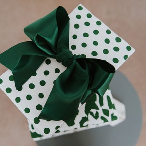 May include: A wrapped gift box with white paper featuring green polka dots. The box is tied with a large, dark green satin bow. The gift box is sitting on a gray surface.