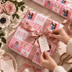 May include: A square gift box wrapped in custom pink paper featuring repeating photos of a girl, the text "ELLIE" and "HAPPY 6TH BIRTHDAY," tied with a dusty rose satin ribbon and a white gift tag reading "Happy Birthday Ellie x ♡."
