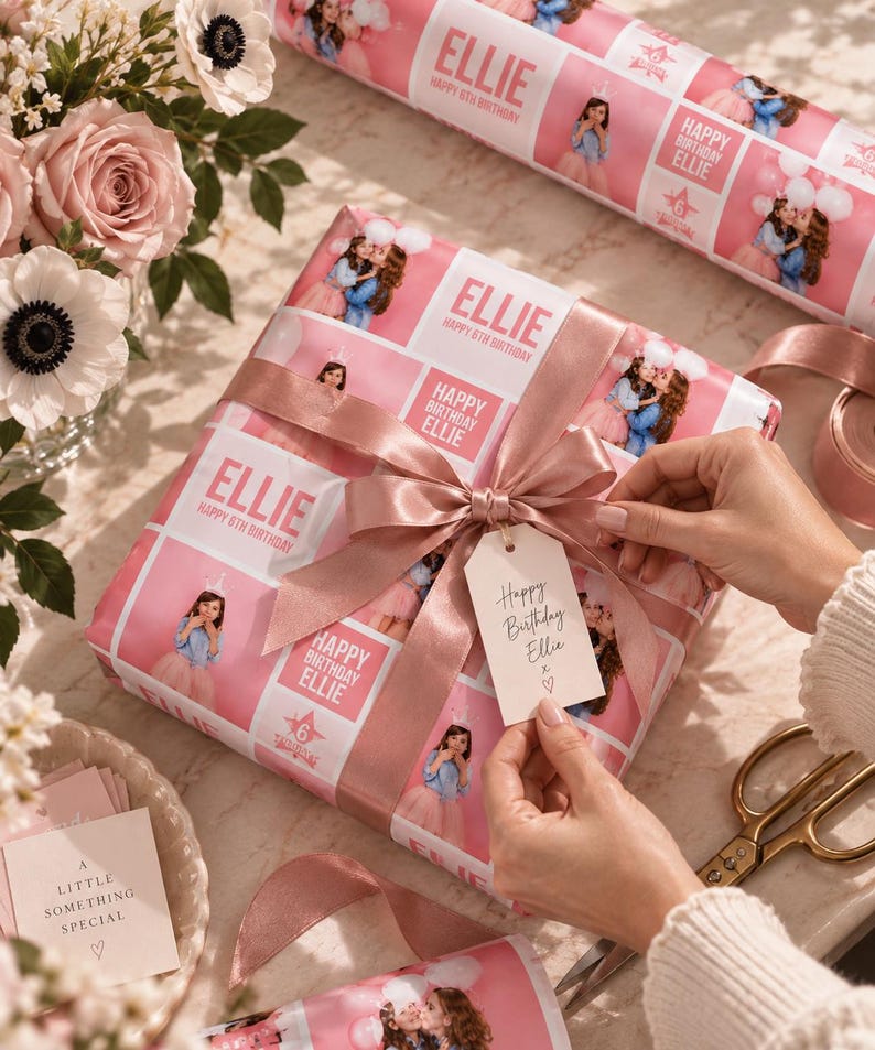 May include: A square gift box wrapped in custom pink paper featuring repeating photos of a girl, the text "ELLIE" and "HAPPY 6TH BIRTHDAY," tied with a dusty rose satin ribbon and a white gift tag reading "Happy Birthday Ellie x ♡."
