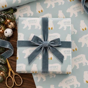 May include: A square gift box wrapped in light blue paper featuring a repeating pattern of white polar bears and the text Arthur's First Christmas. The box is tied with a wide, dark blue velvet ribbon tied in a centered bow.
