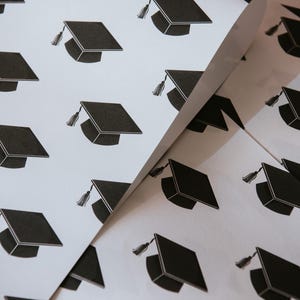 May include: White wrapping paper with a repeating pattern of black graduation caps. The caps have a tassel on the left side.