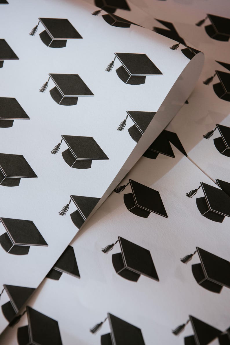 May include: White wrapping paper with a repeating pattern of black graduation caps. The caps have a tassel on the left side.